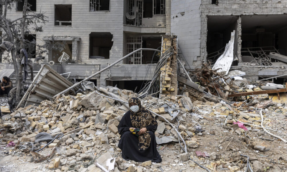 photo of an Iranian woman dressed in black and wearing a face mask sitting in front of a pile of rubble and a bombed-out building in the background