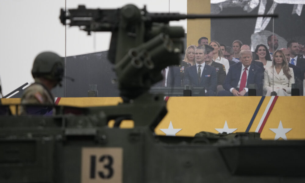 photo of a US military vehicle being driven by a soldier in the foreground, passing by a grandstand where President Trump watches along with his wife, secretary of defense, and others