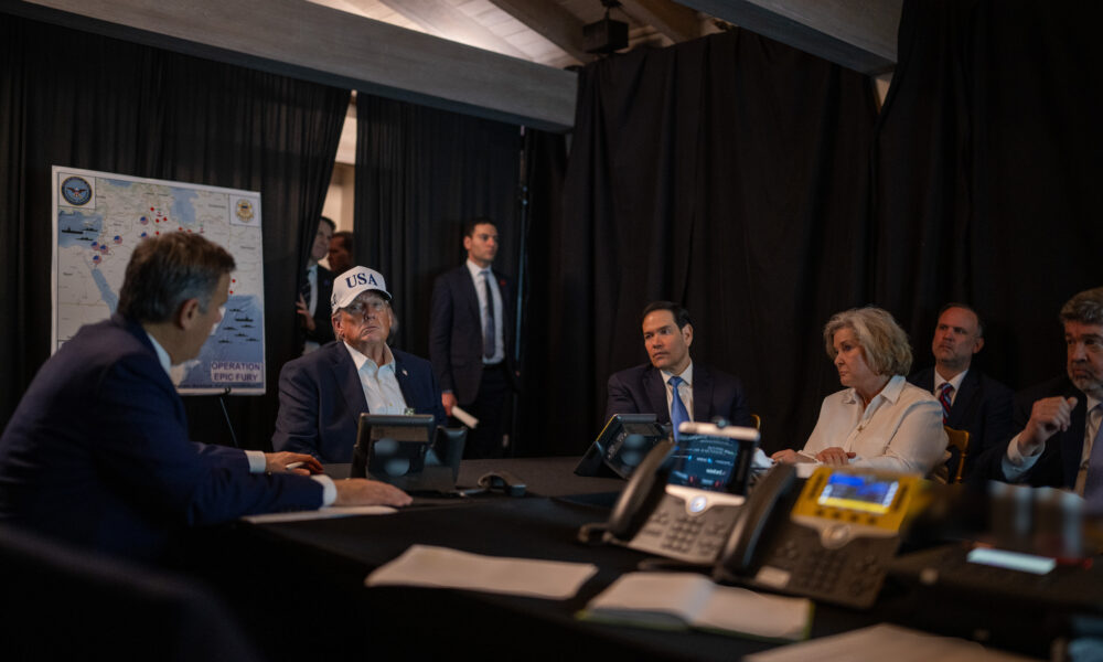 photo of President Trump and members of his cabinet listening to a presentation on the war with Iran in a dark room at Mar-a-Lago; Trump wears a hat with "USA" in large letters, and in the background is a US flag and a screen displaying a map of the Middle East