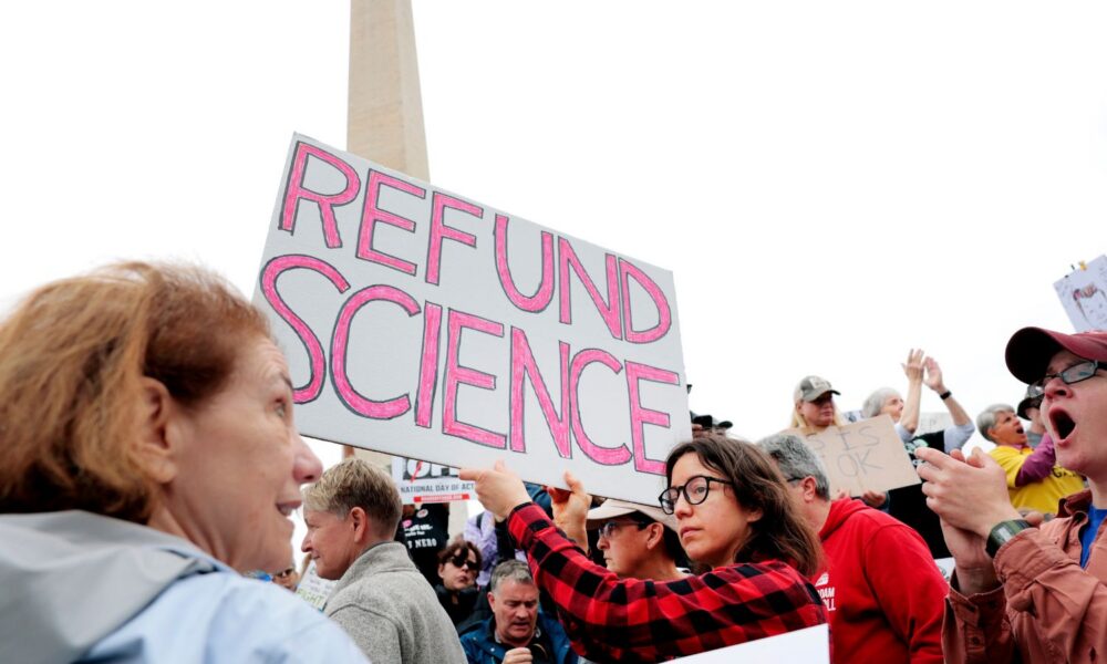 The Washington Monument is in the background of a science-themed protest, with a sign reading REFUND SCIENCE in the foreground.
