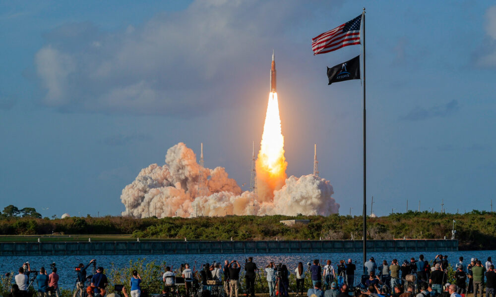 photo of the Artemis II launch; many people are watching in the foreground, across a body of water from the launch site, and a US flag flaps in the breeze