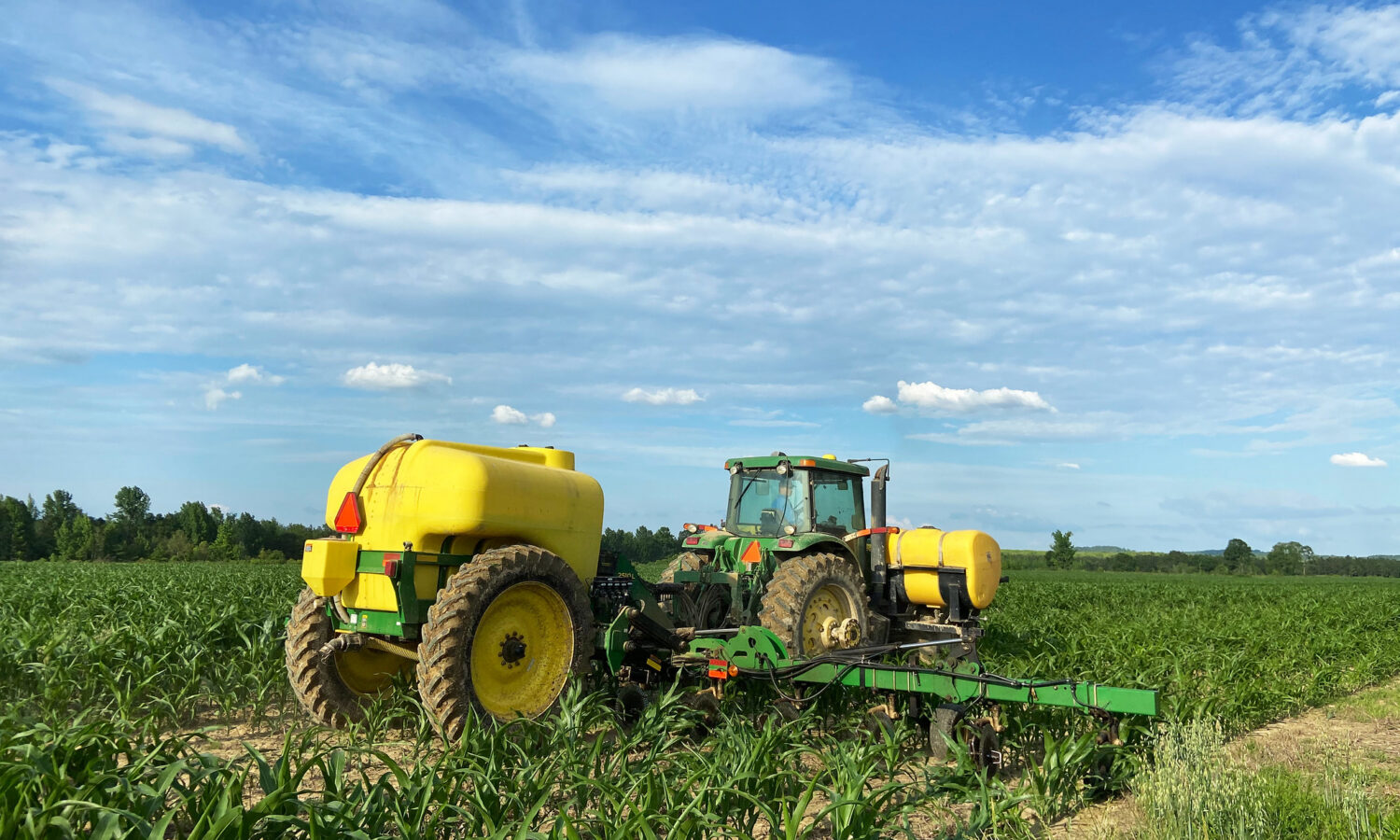 photo of a green tractor pulling a trailer with spraying arms and a yellow tank of fertilizer through a field of young corn plants, under a partly cloudy sky