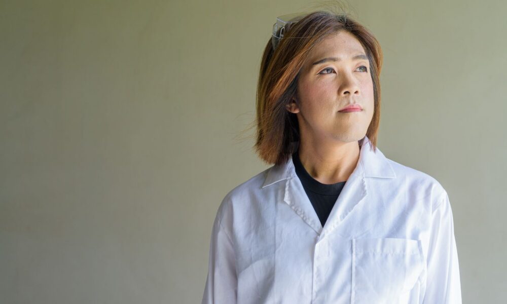 Confident transgender non-binary person in lab coat and protective glasses.