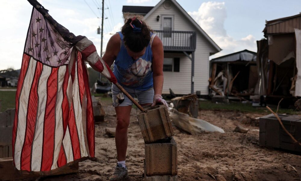 A woman mounts an American flag to a stack of cinderblocks outside of a destroyed mobile home in the aftermath of Hurricane Helene flooding in North Carolina.