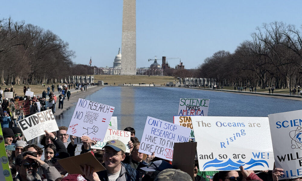 a crowd shot of a protest in Washington DC, with the Washington Monument in the background and attendees holding signs highlighting the importance of scientific research