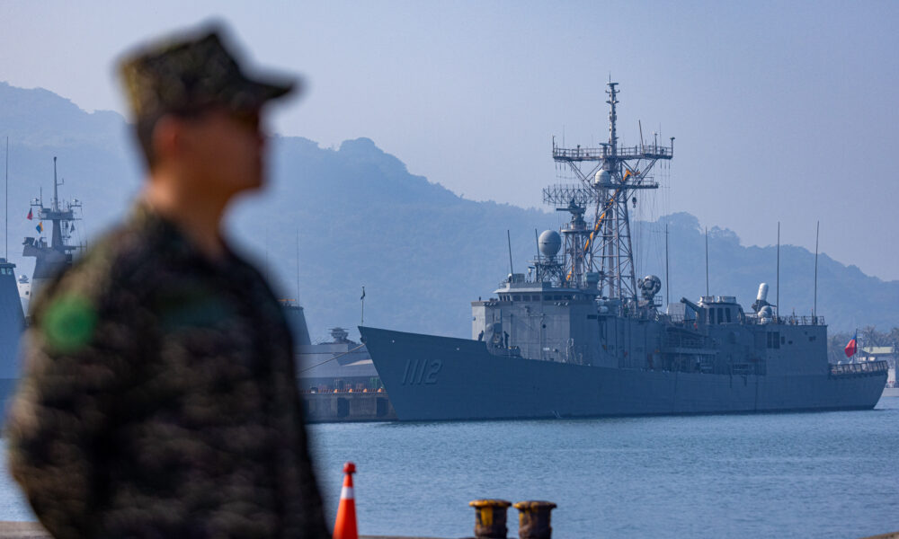 photo of a Taiwanese navy ship docked in a harbor with rocky hills rising up in the background and the blurry image of a soldier standing guard in the foreground
