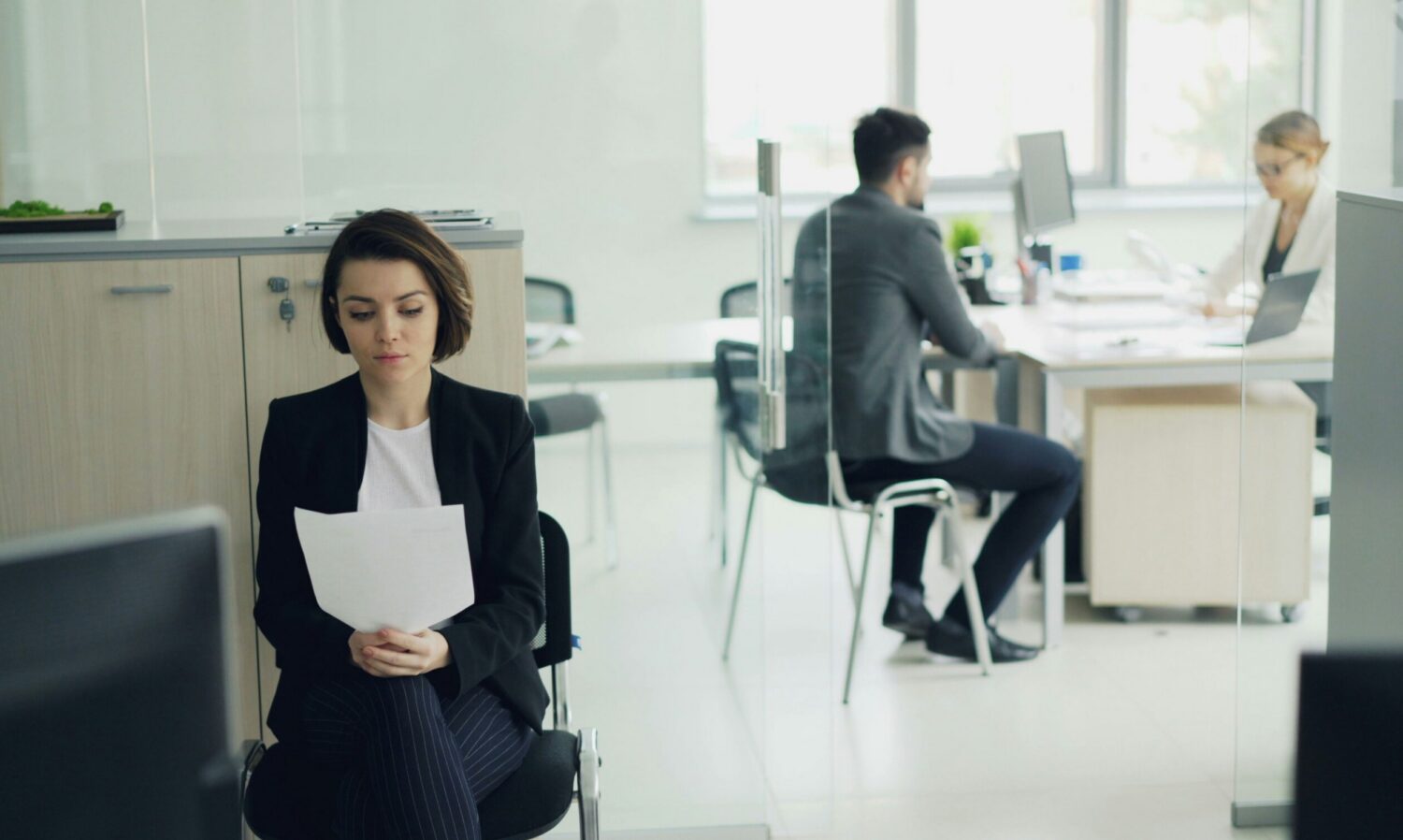a woman sits in an office reading a piece of paper. in the background, two people sit at desks