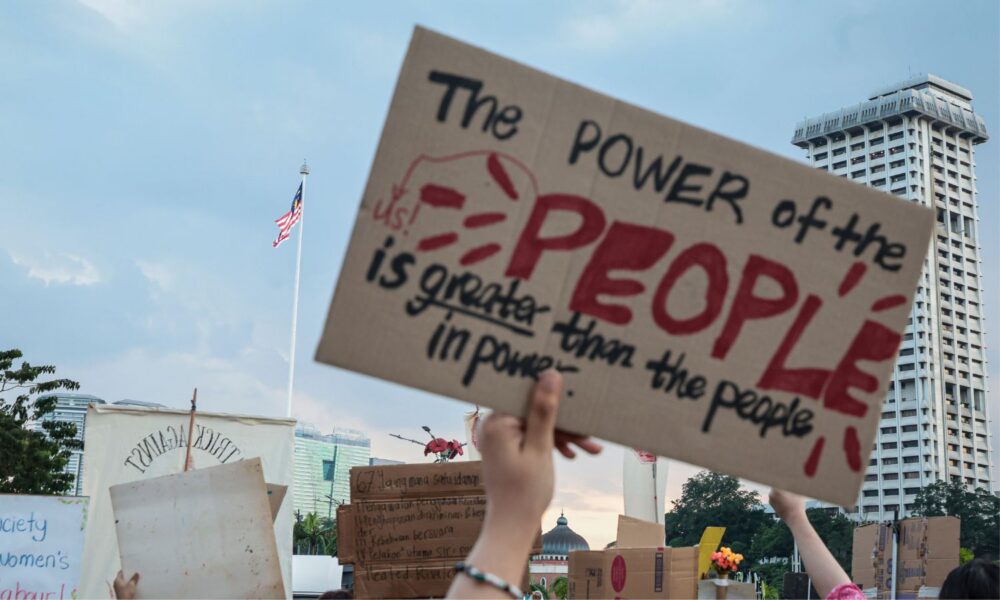 a hand holds a sign reading "the power of the people is greater than the people in power"