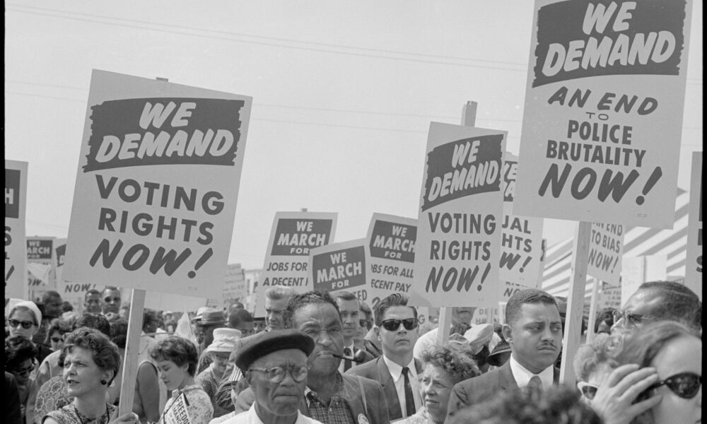 a black-and-white photo of the 1963 March on Washington, featuring a crowd of people holding signs that say "We Demand Voting Rights Now"