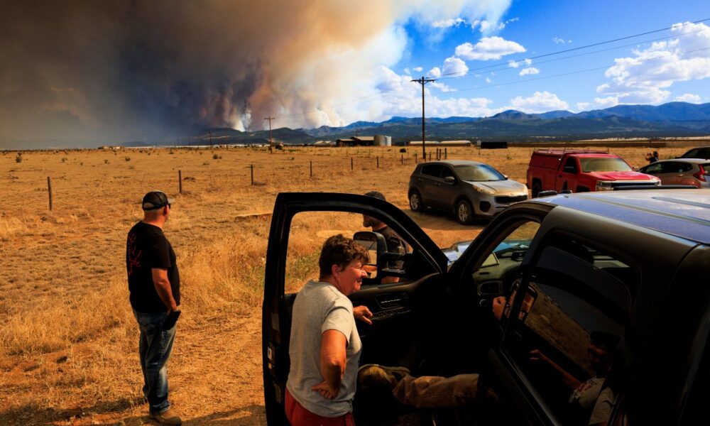 People in Colorado look at the distant Aspen wildfire from a pickup truck. The sky is half blue with clouds, and half orange with billowing smoke from the fire.