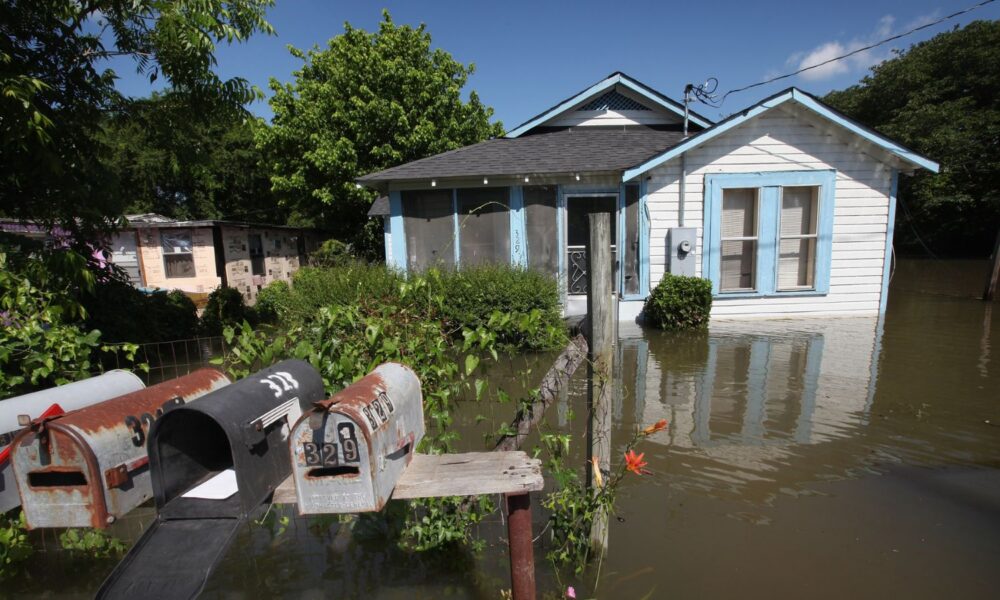 Floodwater surrounds a small white home with blue shutters and multiple mailboxes out front, in Mississippi.