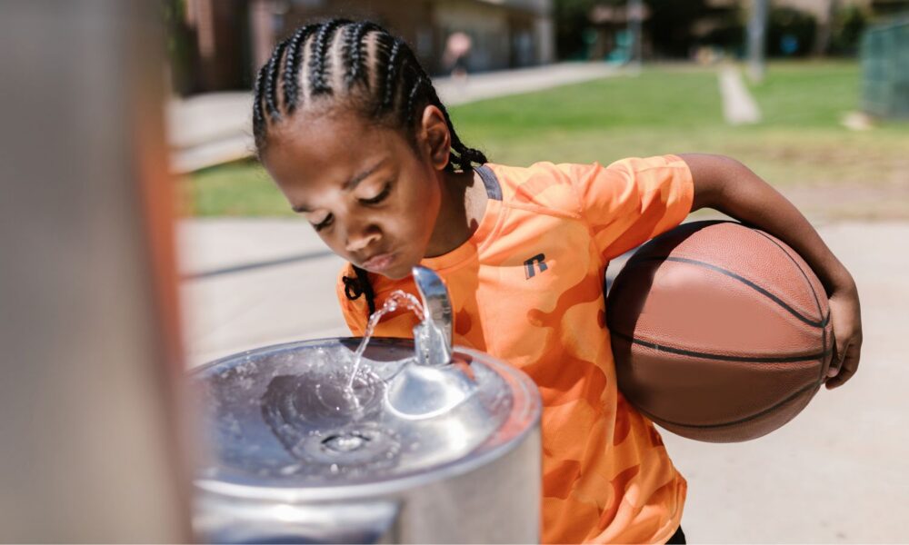 A child in orange holding a basketball drinks from a water fountain