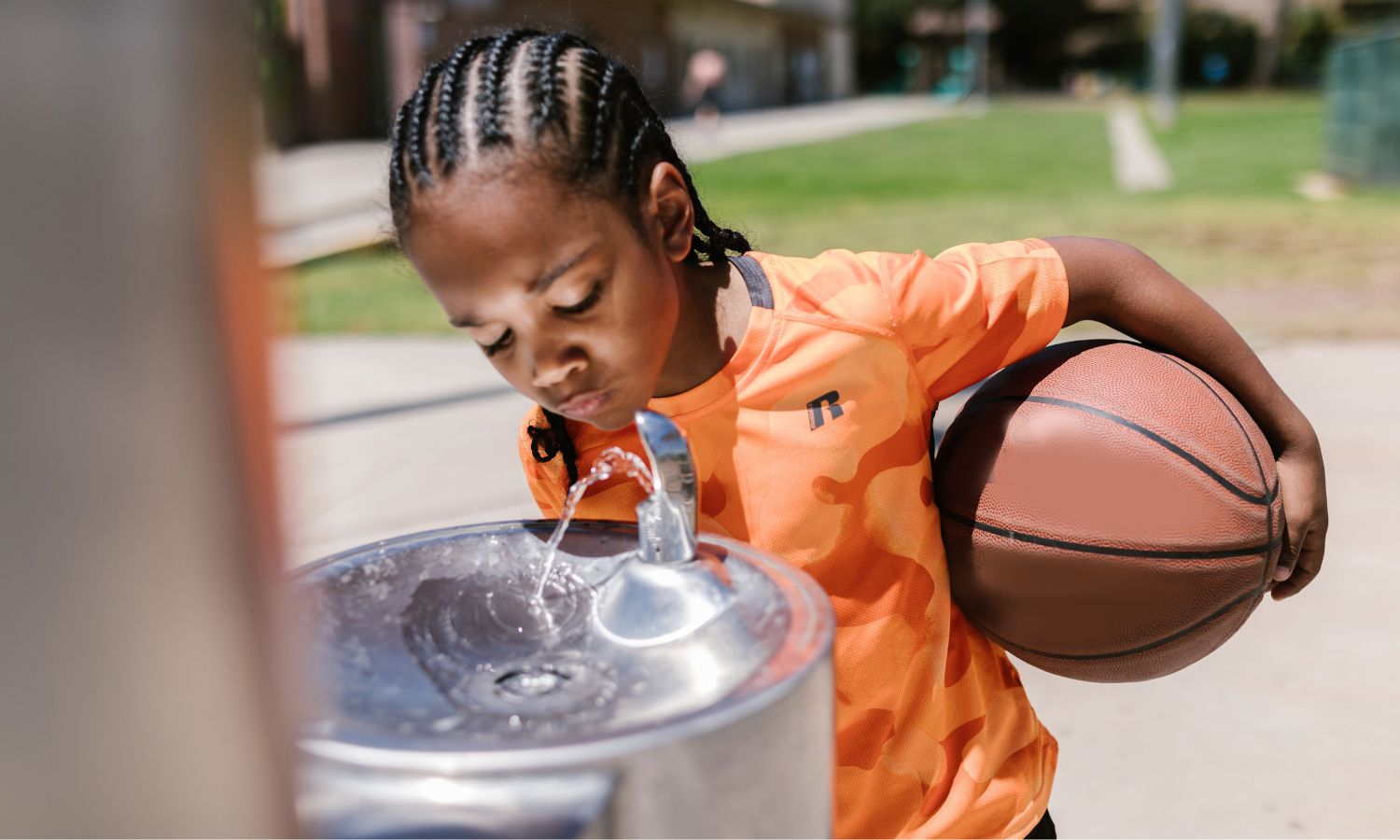 A child in orange holding a basketball drinks from a water fountain