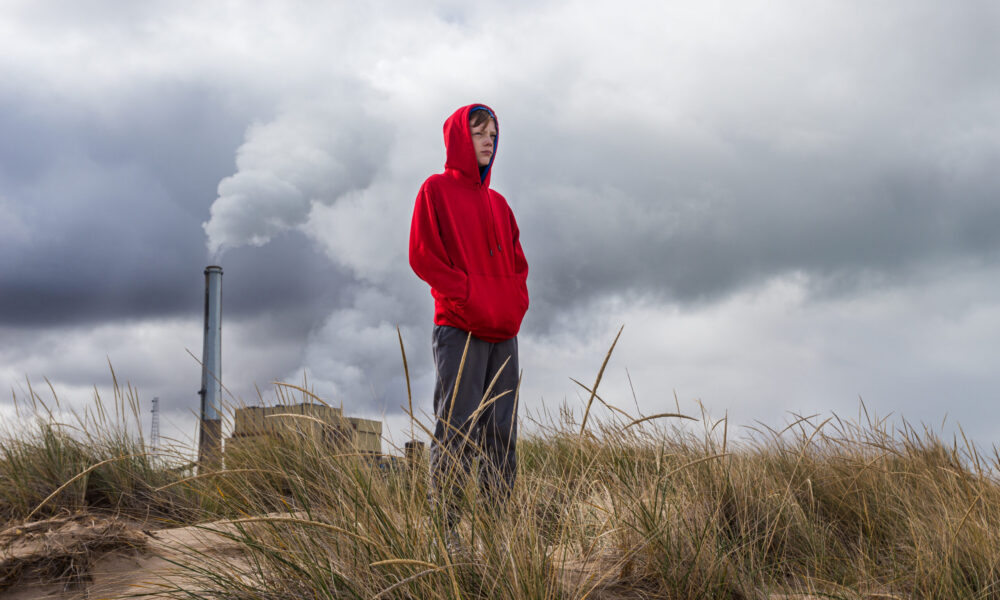 A boy in a red sweatshirt standing in a field with a power plant in the background emitting a cloud